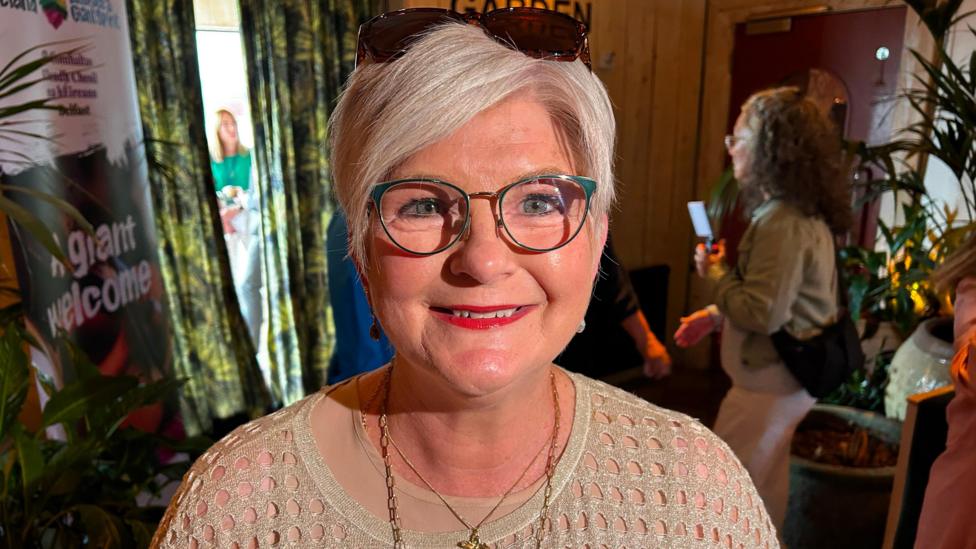 A woman in a light brown jumper wearing necklaces. She has white short hair and wears glasses and red lipstick. There are sunglasses on her head.