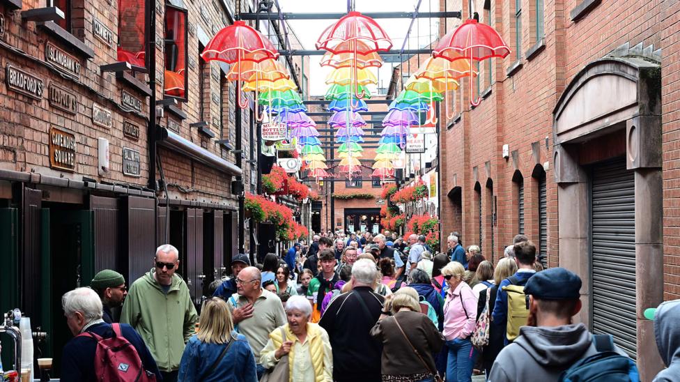 People in the Cathedral Quarter in Belfast under the multi-coloured umbrellas. There are pubs either side.