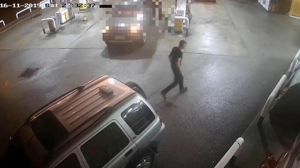 A CCTV camera view shows a person walking across a wet petrol station forecourt at night, with several vehicles parked near the fuel pumps and a timestamp visible in the upper-left corner.