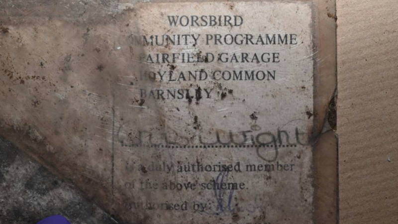 A worn and heavily stained paper card or document inside what appears to be a transparent plastic sleeve. The document is lying on a rough wooden or cardboard surface and is covered with dirt, scratches, dark marks, and signs of age, making much of the text difficult to read. The card contains printed text in black capital letters. The upper section appears to read: WORSBIRD COMMUNITY PROGRAMME AIRFIELD GARAGE HOYLAND COMMON BARNSLEY Some letters are obscured by dirt and damage, so parts of the text are difficult to confirm with complete certainty.