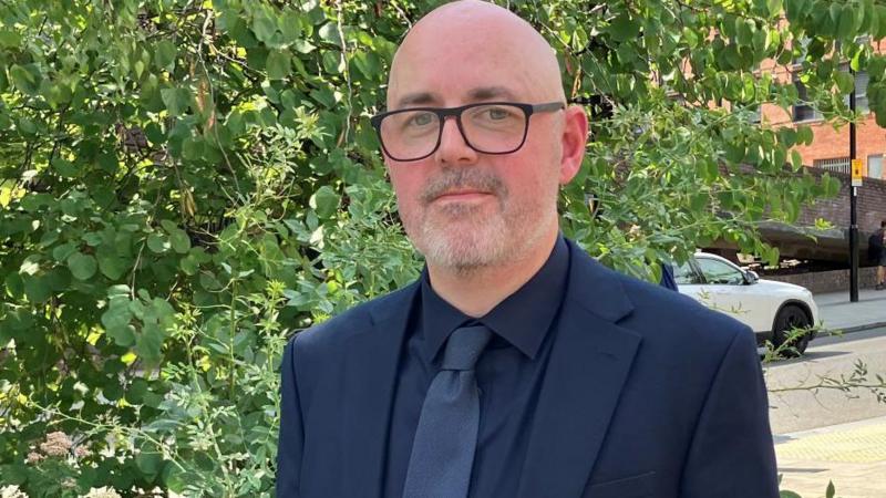 Det Con Ryan wearing a dark suit, dark shirt, and tie stands on a sunlit urban pavement beside leafy greenery, with modern brick buildings, parked cars, and pedestrians visible in the background.