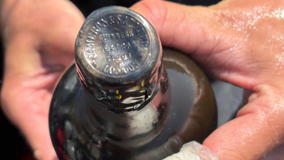 A pair of hands holding a black bottle. Written on the top of the cap is 'GUINNESS EXTRA STOUT LONDON'.