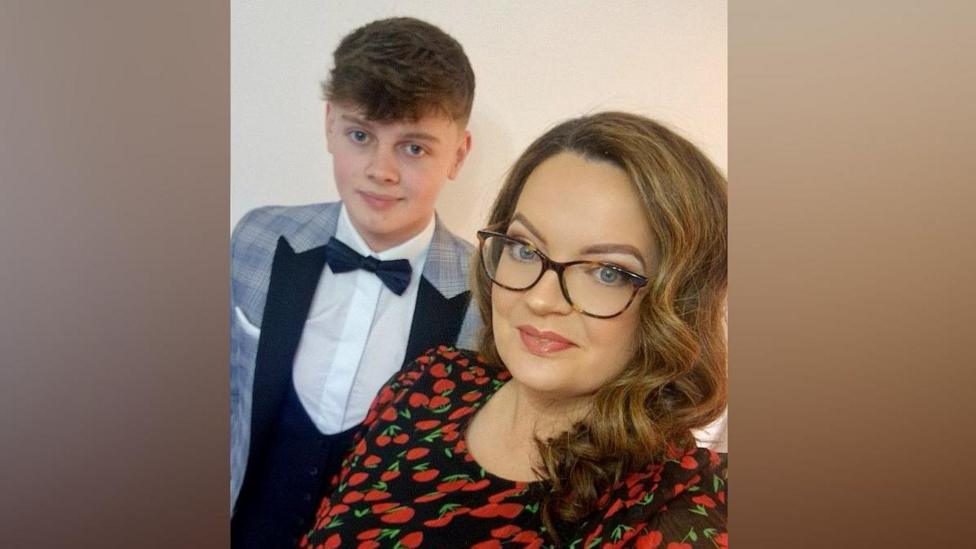 A teenage boy and woman pose for a selfie. He is wearing a suit, dickie bow and while shirt, and blue checked jacket. To his left a woman stands holding the phone to take the picture. She is wearing a red dress and has brown shoulder length hair