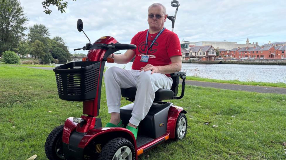 Stephen Magee sitting on his mobility scooter, which is red. He is wearing a red T-shirt with light grey jogging bottoms and lime green crocs. The scooter is parked on grass in a park beside the River Bann which can be seen behind him.