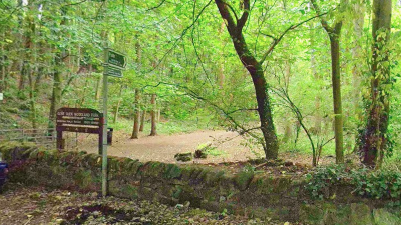 A park entrance with a wooden sign that reads Gore Glen Woodland Park with a pathway surrounded by green trees