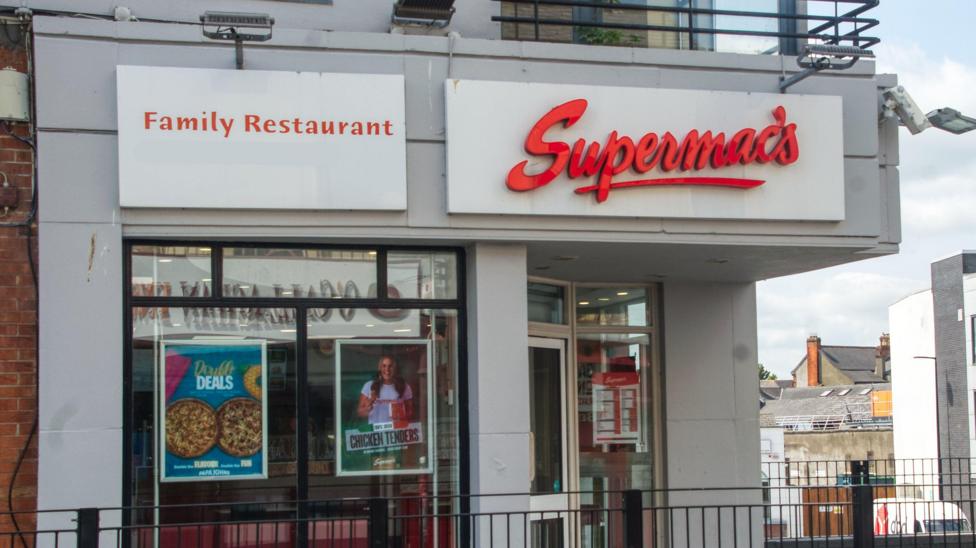 The exterior of a Supermac's fast food restaurant in Ireland. A large red letter sign shows the name of the business above a door and glass fronted store