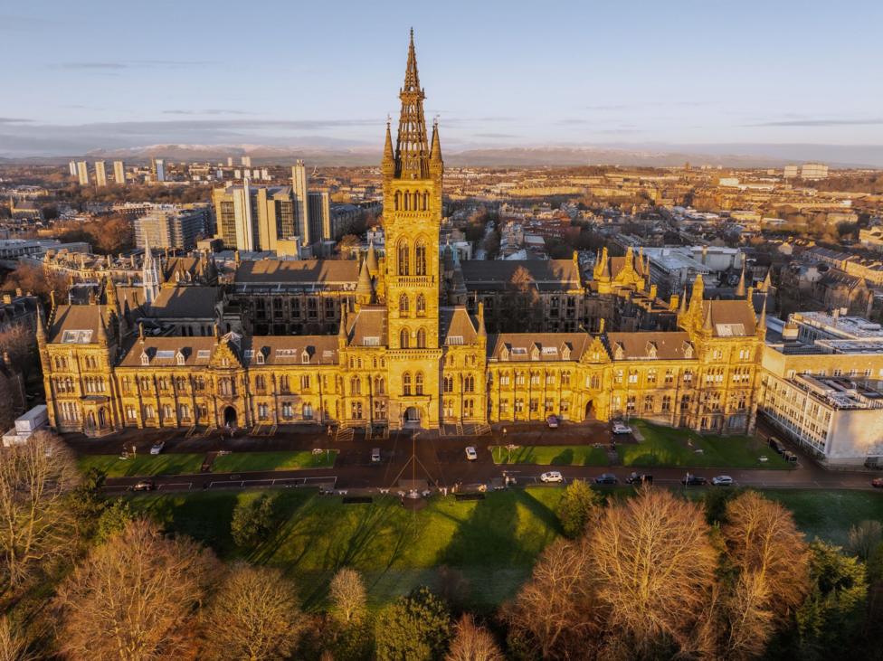 Aerial view of the University of Glasgow's main Gilbert Scott Building, with its prominent central tower and Gothic architecture, overlooking the surrounding city skyline in low winter sunlight.
