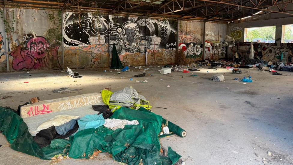 Interior of a large abandoned warehouse with graffiti-covered walls and exposed metal roof beams. The concrete floor is scattered with rubbish, clothing, and discarded materials. In the foreground, torn green tarpaulins and fabric lie beside a low concrete structure with graffiti. Large black-and-white mural artwork covers part of the back wall, while daylight enters through windows on the right side of the building.
