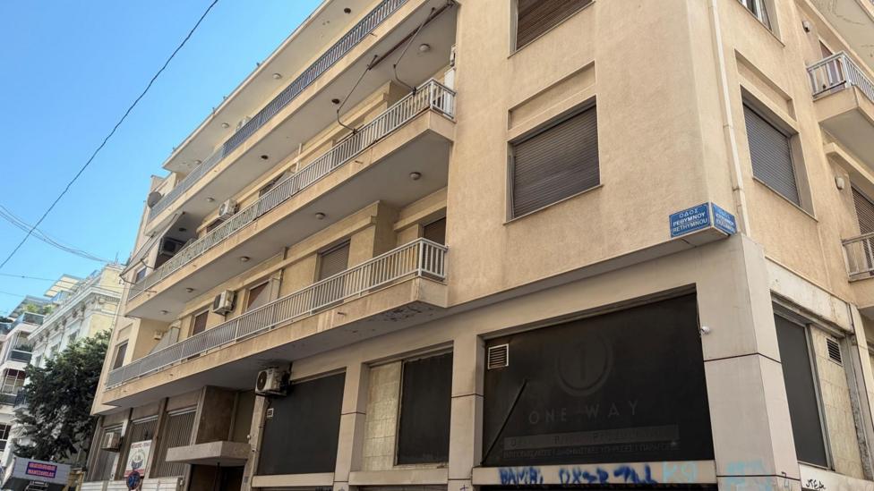 9 Rethymnou Street in Athens, where Elisabeth-Jane Ross was staying before she was killed - Exterior view of a multi-storey beige apartment and commercial building on a street corner. The building features several balconies with metal railings, shuttered windows, and air-conditioning units. At street level, large dark shopfront windows display faded signage and graffiti. A blue street sign with Greek text is mounted on the corner of the building, and the scene is lit by bright daylight under a clear blue sky.