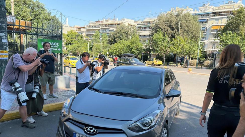 Several photographers standing on a city street photograph a silver Hyundai car parked at the roadside. In the foreground, camera operators with large telephoto lenses aim toward the vehicle, while a person in a dark uniform stands nearby. Trees, apartment buildings, yellow taxis, and Greek-language street signage are visible in the background under clear daytime skies.