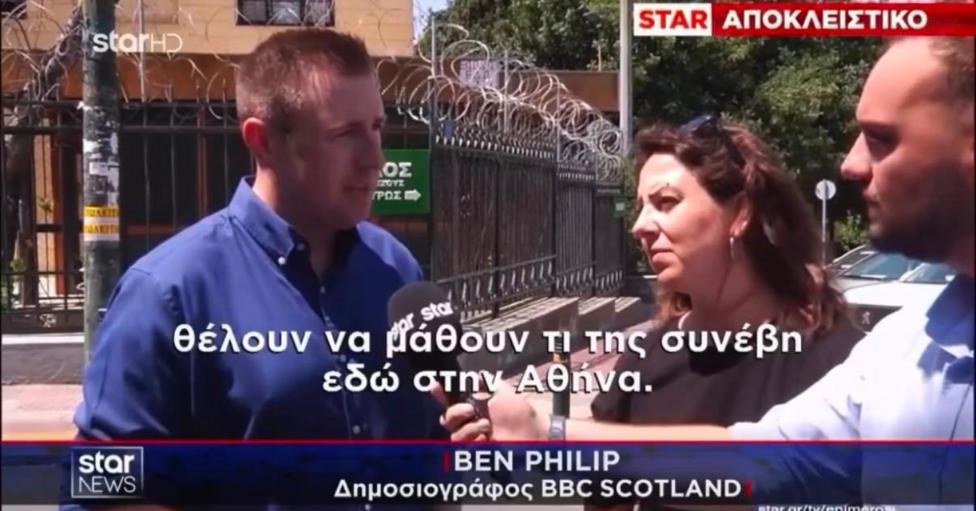 Television news screenshot showing an outdoor interview beside a metal security fence topped with razor wire. Three people are visible in the foreground, with one person speaking into a microphone while another holds recording equipment. Greek-language captions appear across the screen, along with a news banner and the text
