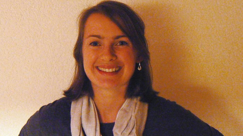 Head-and-shoulders portrait of a person standing indoors against a plain cream-colored wall. The person has shoulder-length dark hair, is wearing a dark top, a light-colored scarf, and a small drop earring, and is facing the camera. The image is lit with warm indoor lighting and has a simple, uncluttered background.