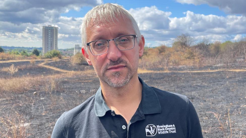 A man with white-grey hair and black glasses. He is wearing a black t-shirt that has a badger logo on the breast and says