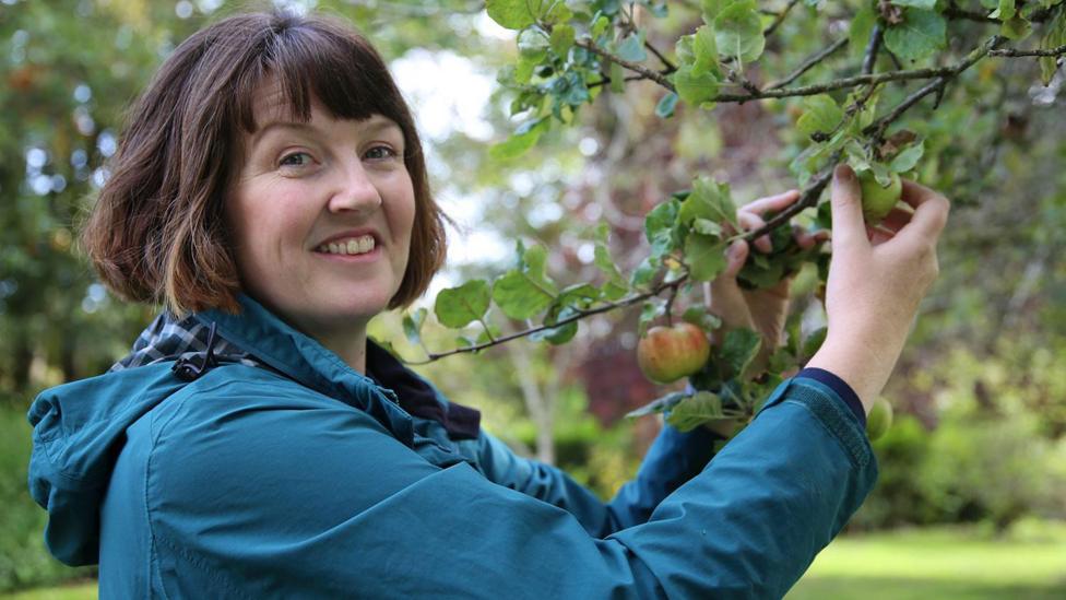 A woman with short dark hair, wearing a blue jacket, holds an apple tree branch and smiles at the camera. She is pictured side-on, from the shoulders up, with an orchard in the background, blurred.