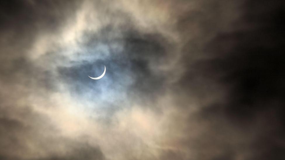 A partial solar eclipse in a cloudy night sky. A crescent of white moon can be seen.