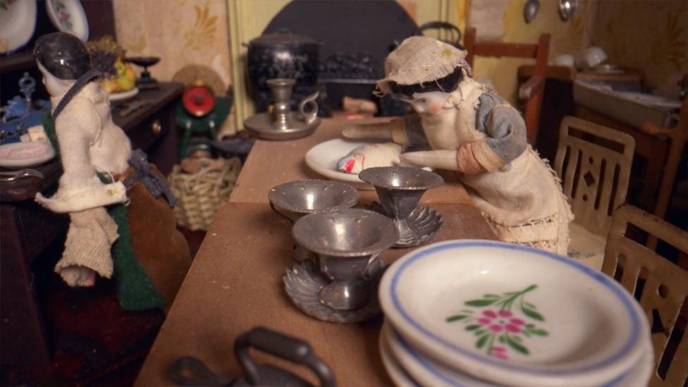 The inside of a doll house with a small miniature figure in a pinafore preparing fish on a plate at a table which is covered with metal cups and white flowered plates.