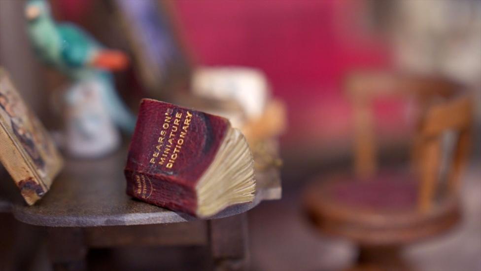 A table in a doll house with a burgundy and gold lettered book saying Pearson's miniature dictionary on it. The book sits on a grey table with a dark wooden chair with a burgundy cushion in the background which is blurred out.
