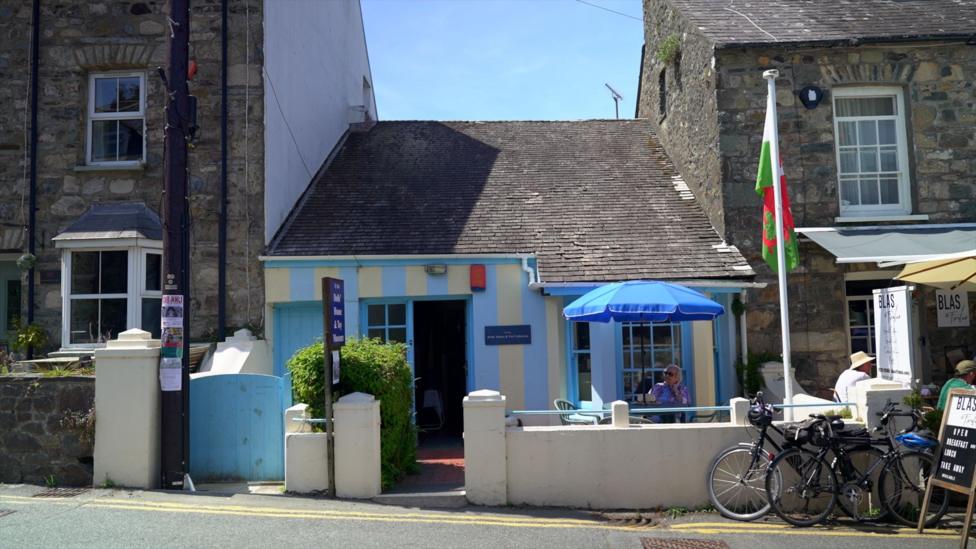 The view from the other side of the street of the single story dolls house museum. It sits with a grey slate roof sandwiched between two grey stone brick houses. The outside of the museum has been painted with a thick white and blue stripe pattern and a table with a blue parasol is positioned outside beside a welsh flag pole.