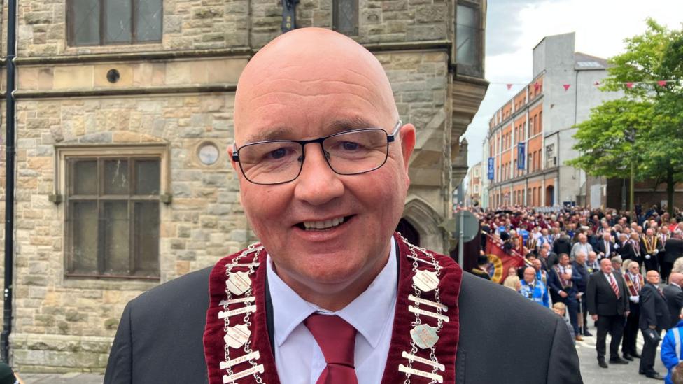 William Walker is smiling and wearing glasses. He is wearing a red collarette and a grey suit with a red tie.