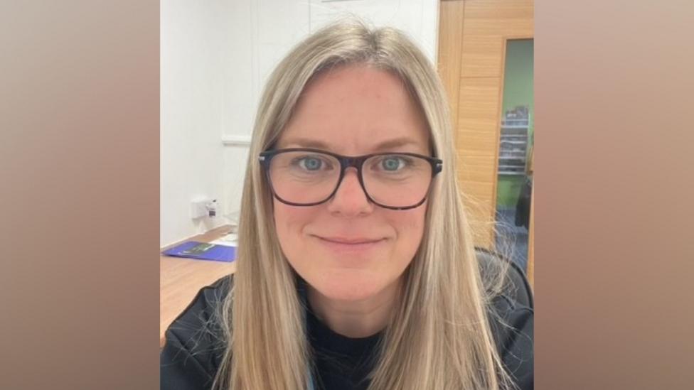 A woman with blonde hair. She has big glasses and is wearing a black t shirt.
