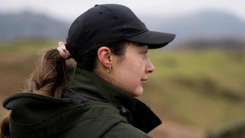 The side profile of Jessie Roper who is looking to the right. She is wearing a big green coat with a hood and a black cap. Her hair is tied in a plait with a pink scrunchie and she is wearing a gold hoop earring. She has a straight face and is standing outside with hills in the background which is out of focus.
