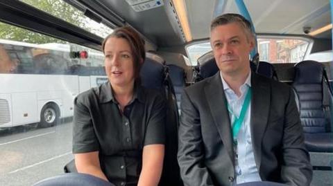 Transport Minister Liz Kimmins, a woman with dark hair tied back, pictured sitting with Translink Chief Executive Gerard Carlin, a man with short, grey hair, as the travel on one of the Fleadh shuttle bus services. She is wearing a black short-sleeved shirt and he is wearing a grey suit and a white shirt.