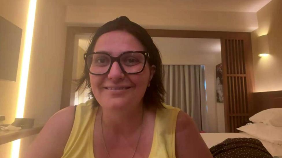 A smiling Hayley Chapman is sitting in a hotel room with a yellow vest top and glasses.