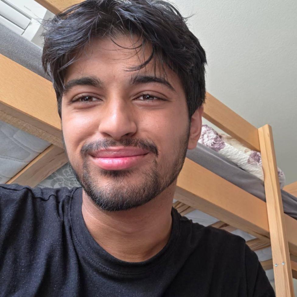 A man with black hair and a black beard smiling, he is wearing a black top and behind him is wooden bunk beds. 
