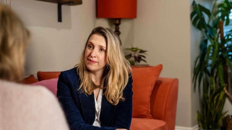 A woman with long blonde hair sat on an orange sofa, she is wearing a dark blue blazer and white blouse and is in conversation with a woman whose face is not visible, she has brown hair and is wearing a beige jumper. 