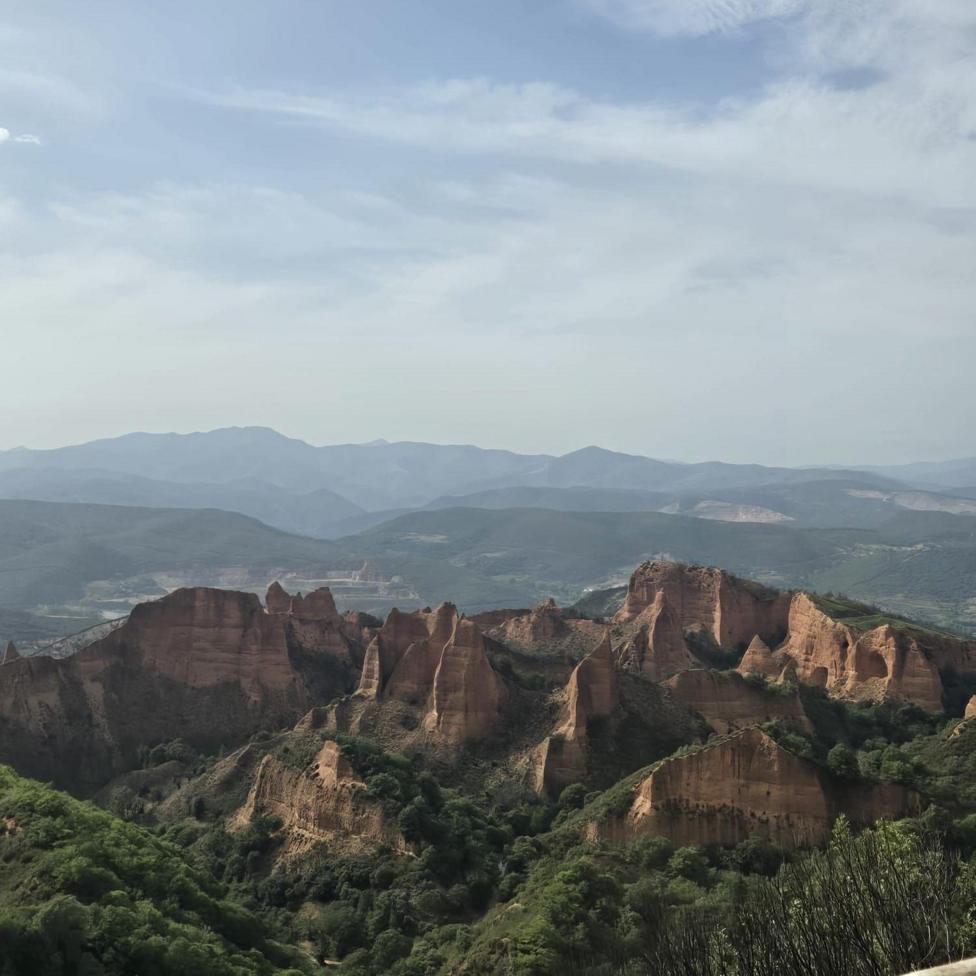 Rock formations with steep, vertical cliffs and pinnacles dominate the foreground of a wide valley landscape. Areas of green vegetation surround the formations, with layered hills and mountains extending into the distance. Thin cloud cover fills much of the sky above the ridge line.