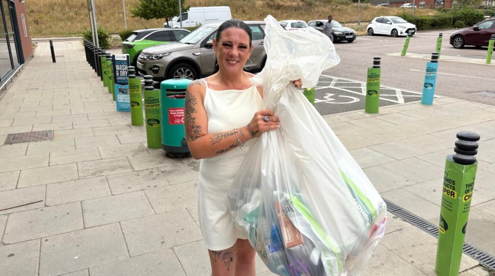 Sandra McCluskey standing on a pavement outside a supermarket holds a large transparent sack filled with recyclable items, including plastic bottles, cartons and packaging. She is wearing a light-coloured sleeveless outfit and has visible tattoos on both arms and one leg. Behind her are recycling bins, a row of green and blue bollards, parked cars and a supermarket car park. The photo was taken outdoors in daylight, with dry grass visible on a bank in the background.