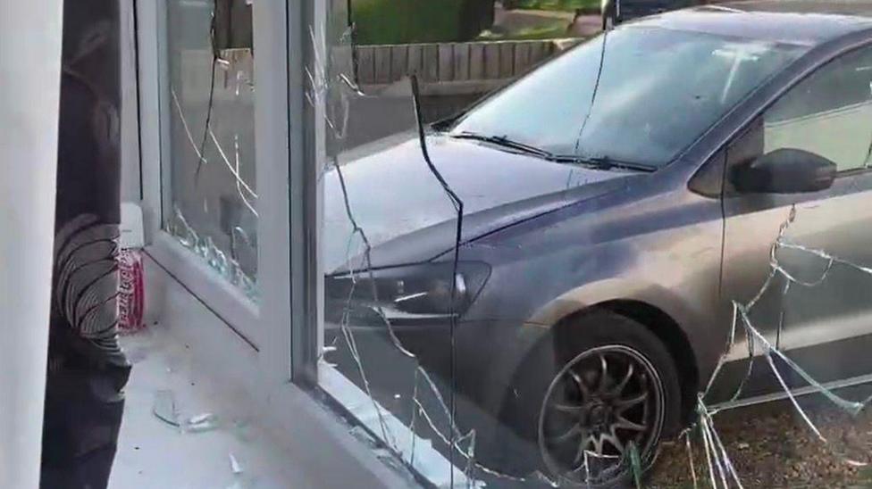 View from inside a house looking out through a shattered front window. The glass pane is covered in large cracks and several sections are broken, with shards of glass scattered across the white windowsill. Outside, a grey car is parked close to the window on a residential street lined with trees and hedges. The image appears to document damage to the property, with the broken window as the main focus.