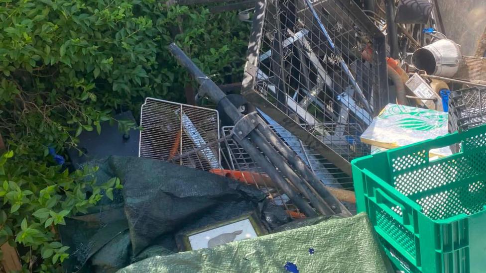 A large pile of dumped waste and scrap materials stacked beside a hedge and a dark vehicle. The debris includes metal fencing panels, poles, crates, tools, tarpaulins and assorted household items, with vegetation partially growing around the rubbish. The scene appears to show fly-tipped waste in an overgrown outdoor area on a sunny day.