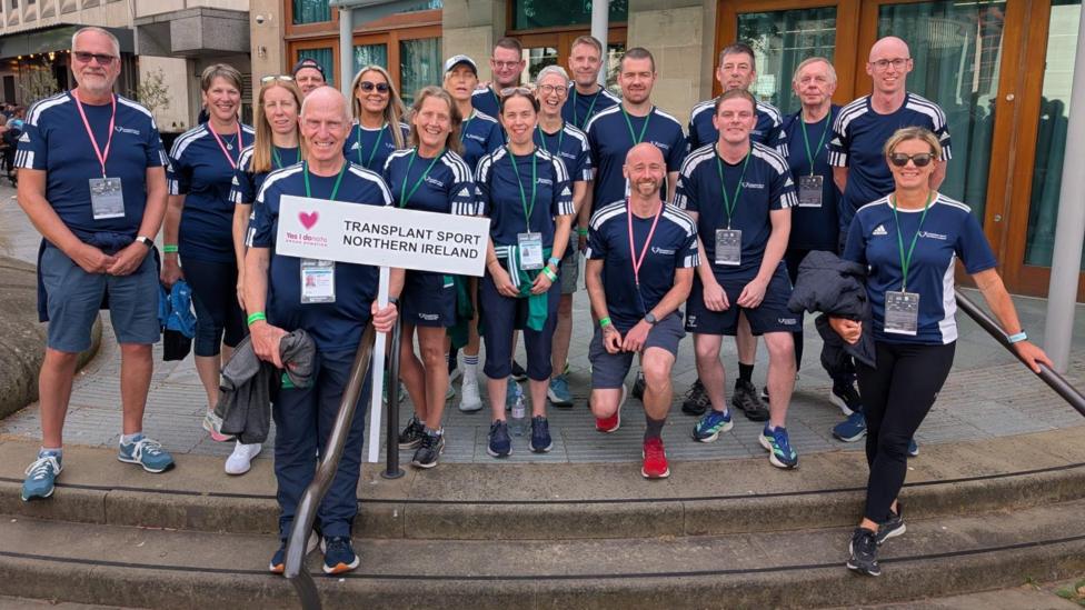 A team photo of Transplant Sport Northern Ireland. They are all standing on concrete steps. One man near the front is holding a white sign reading: 'Transplant Sport Northern Ireland'