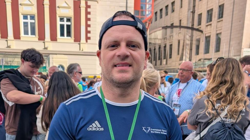 Richie Sheerin is wearing a navy sports T-shirt and a backwards navy cap. He is also wearing a green lanyard. He is standing in front of a large crowd.