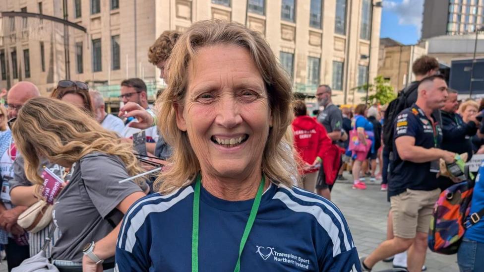 Kay Hack has long blonde hair and is wearing a navy T-shirt and green lanyard. She is standing in front of a large crowd of people.
