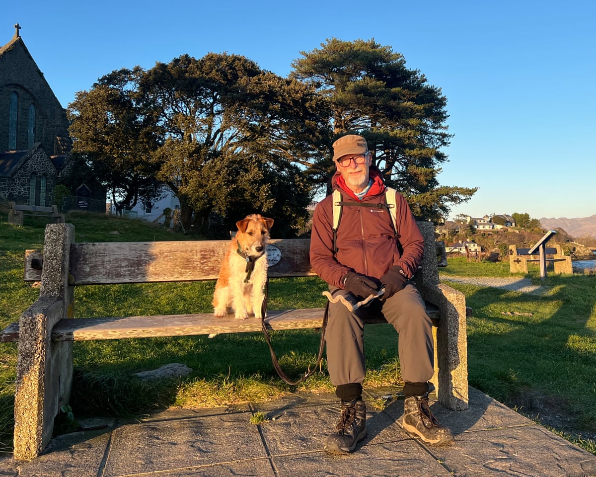 Man in hiking clothes with dog on a public bench. River in background and steep hills