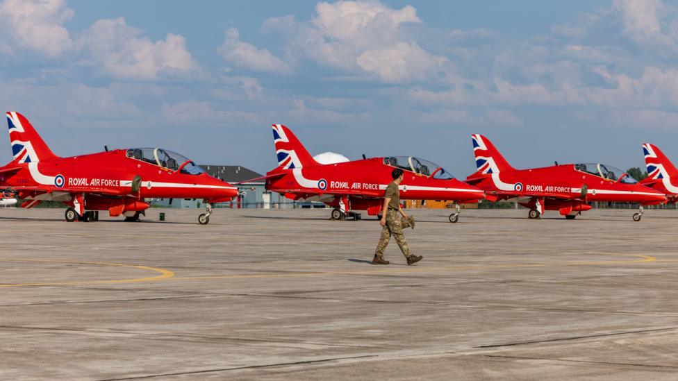 The Red Arrows pictured on the tarmac at Goose Bay in Canada. A person in uniform is walking past the jets.