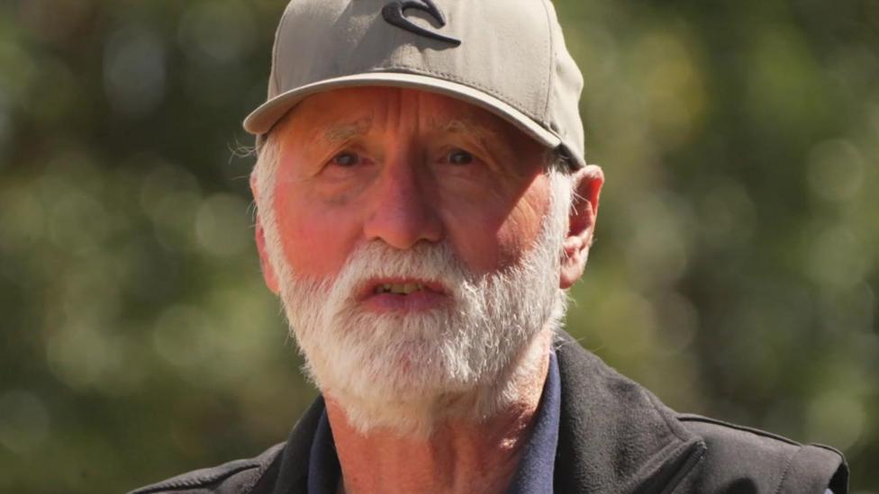 Nigel has a white beard and is wearing a grey cap and dark jacket.