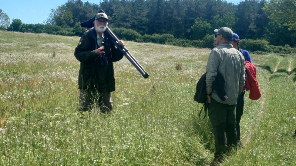 Nigel is standing in a field of tall grass and talking to several people standing on a grassy path. There are trees in the background.