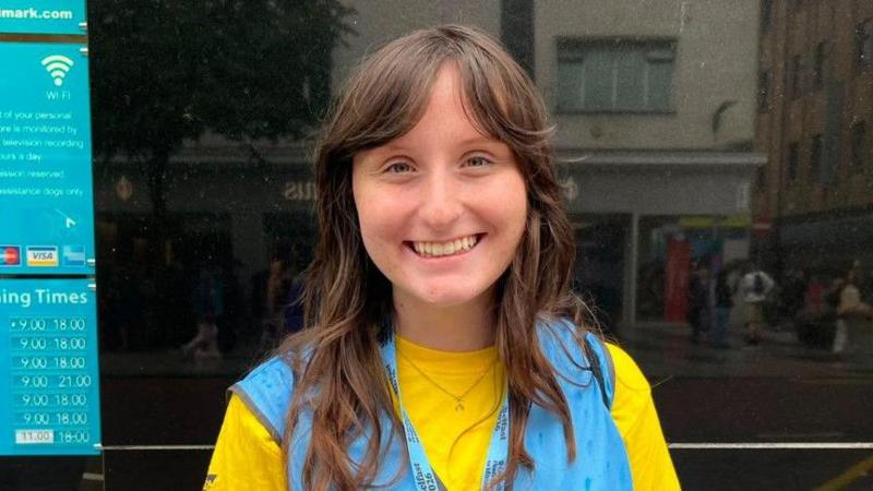A young woman, Addyson McGowan pictured outside Primark home on Royal Avenue. She is wearing a yellow t-shirt with a blue bib on top and a blue lanyard. She has brunette hair.