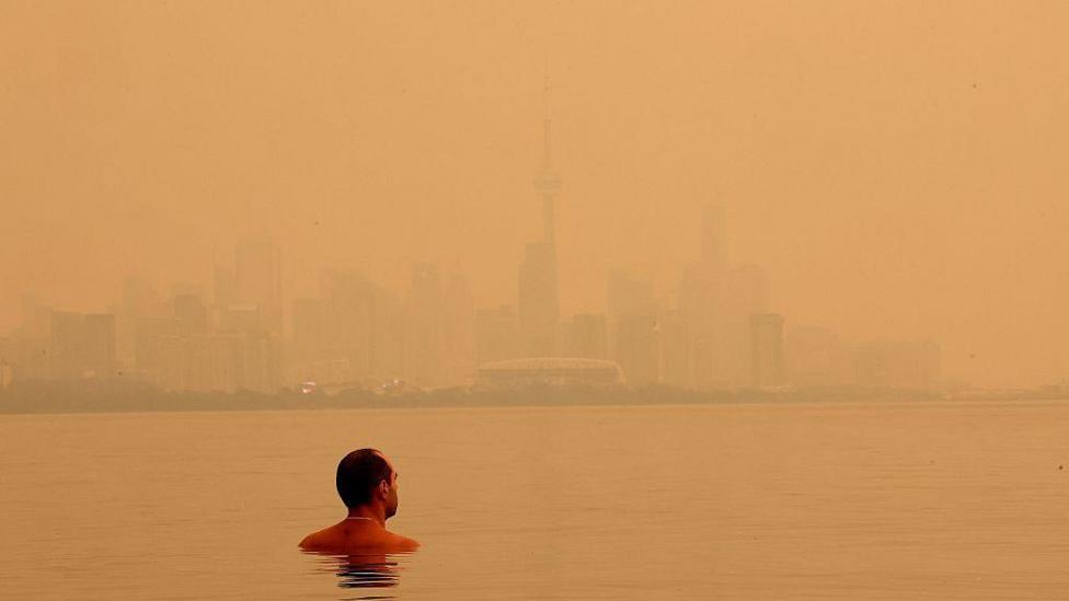 A man swimming in Lake Ontario on 15 July overlooking the Toronto skyline blanketed by a bright, orange haze from wildfire smoke.