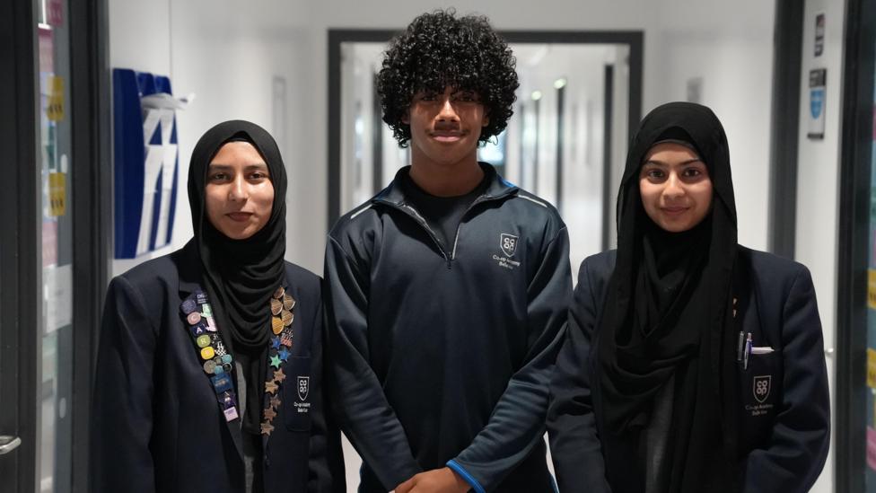 Three pupils stand side by side in a school corridor. Two are wearing dark blazers with a school badge, while the pupil in the centre is wearing a dark sports-style quarter-zip top.