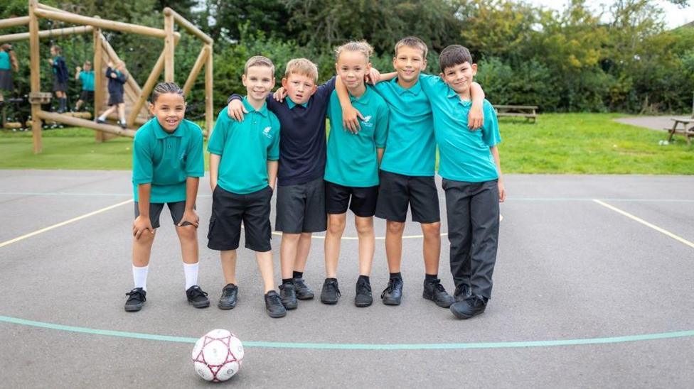 Six primary school pupils stand together on a playground with their arms around each other. Most are wearing turquoise polo shirts and dark shorts or trousers, with a football resting on the ground in front of them and a climbing frame behind.