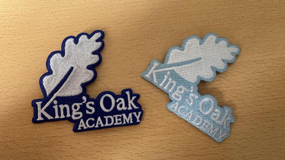 Two embroidered iron-on school badges lie on a light wooden surface. Both feature the words “King’s Oak Academy” beneath a large oak leaf, with one badge outlined in dark blue and the other in a pale blue colour.