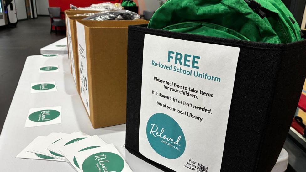 Boxes filled with donated school uniform items are laid out on a table indoors. A sign on the nearest container advertises free