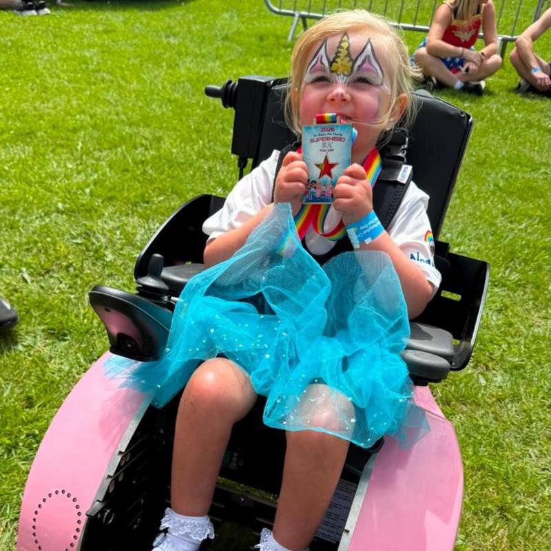 A young blonde girl with a unicorn design painted on her face. She is holding up a medal which is round her neck. She is wearing a white t shirt and blue tutu-style skirt and is sat in a black wheelchair with black straps holding her in. The wheelchair is on a grassy field, with other people sat on the field behind her.