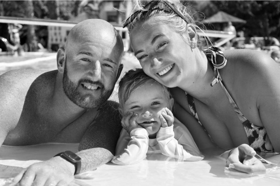 A black and white photo of a man and a woman with a youn girl between them. They are in a row, leaning up on the side of a swimming pool. The man is bald, with a beard, and is shirtless. The woman has ligth hair piled on her head with sunglasses perched on top. She is wearing a swimsuit. The little girls is resting her chin in her hands with her elbows propped up on the side. They are all smiling and looking at the camera.