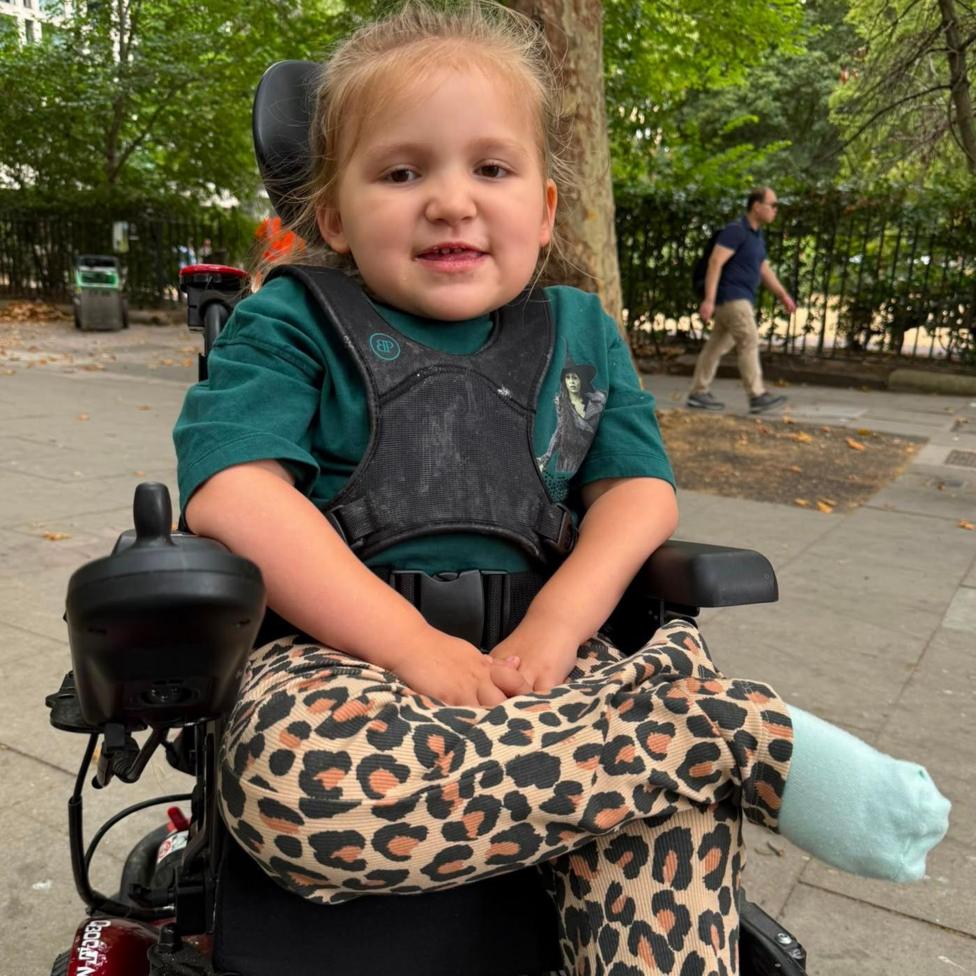 A young blonde girl in a black wheelchair with black straps across her chest. She is wearing a green Wicked t shirt and leopard print trousers. Behind her is a pavement and green-leaved trees.