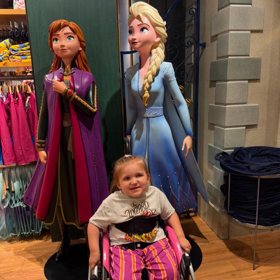 A young blonde girl in a black wheelchair with black straps across her chest. She is wearing a white Wizard of Oz t shirt and pink and orange striped trousers. Behind her are life-size figures of Disney pricesses Anna and Elsa.